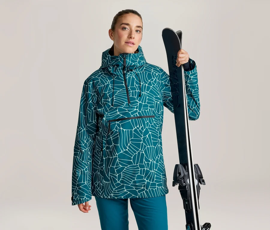 Frau in einem türkisfarbenen Anorak mit weißem geometrischem Muster und türkisfarbener Hose, die Skier über der Schulter hält.