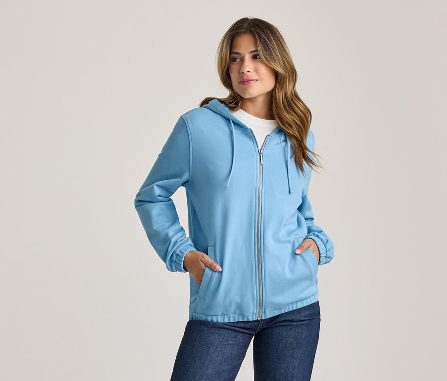 Frau in hellblauer Kapuzenjacke und Jeans, die Hände in den Taschen, blickt nach rechts.