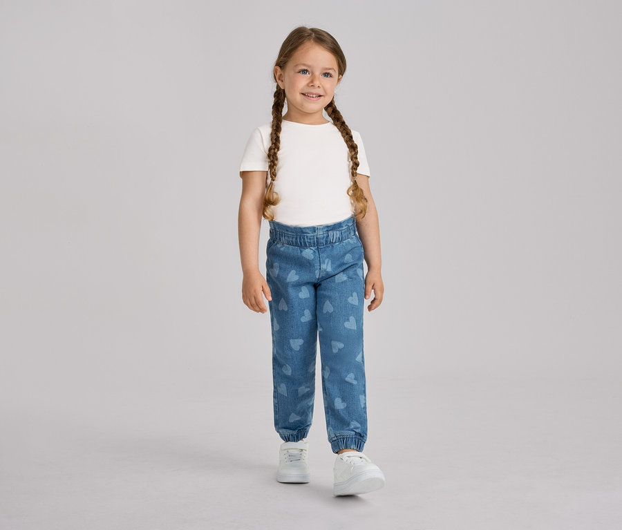 Ein junges Mädchen mit zwei Zöpfen steht lächelnd da und trägt ein weißes T-Shirt sowie blaue Kinder-Pull-on-Pants – Fit »Ella« mit Herzmustern und weiße Turnschuhe.