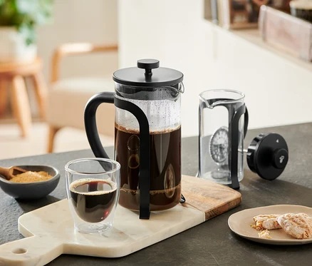 Eine schwarze Siebstempelkanne 800 ml, 6 Tassen, gefüllt mit Kaffee, steht auf einem Marmorbrett neben einem Glas Kaffee, einer Zuckerschale und Keksen. Eine weitere Siebstempelkanne ist zerlegt daneben.