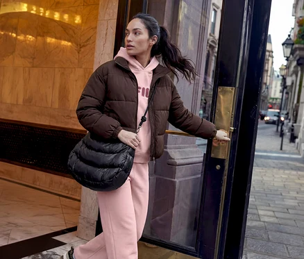 Eine Frau mit dunklem Pferdeschwanz öffnet eine dunkle Tür und blickt auf eine Straße. Sie trägt eine braune Steppjacke, einen pinken Hoodie mit FILA-Logo, eine pinke Jogginghose und eine schwarze gesteppte Umhängetasche.