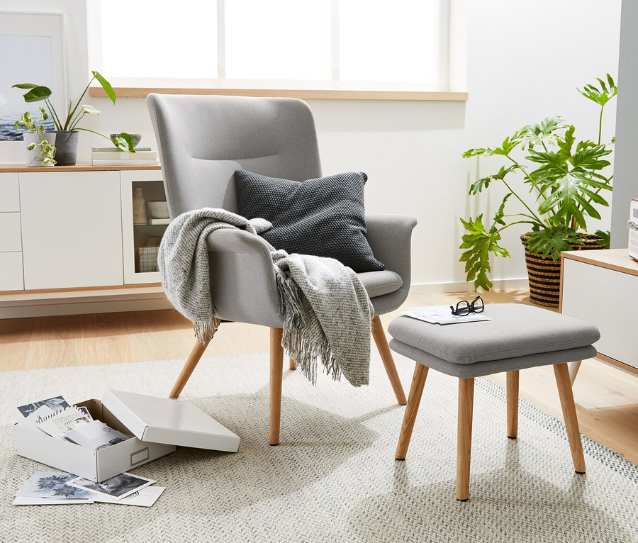 Ein grauer Sessel mit Fußhocker steht im Wohnzimmer neben einer weißen Kommode.