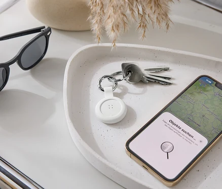 Auf einer Ablage liegen eine Sonnenbrille, Schlüssel, ein weißer Tracker und ein Smartphone mit Karten-App.