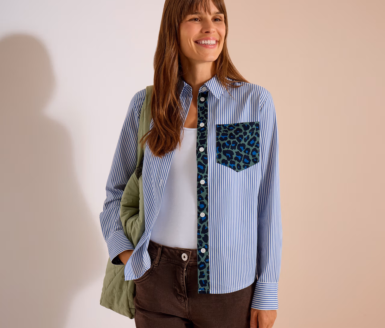 Lächelnde Frau in einer blau-weiß gestreiften CECIL Hemdbluse mit grün-blauem Leopardenmuster an Knopfleiste und Tasche, weißem Top und brauner Hose, mit grüner Stepptasche.