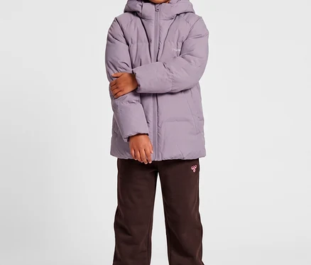 Ein junges Mädchen steht mit verschränkten Armen und trägt eine mauvefarbene hummel® »HMLJR All Weather Puffer Jacket«, braune Jogginghose und weiße Sneaker.