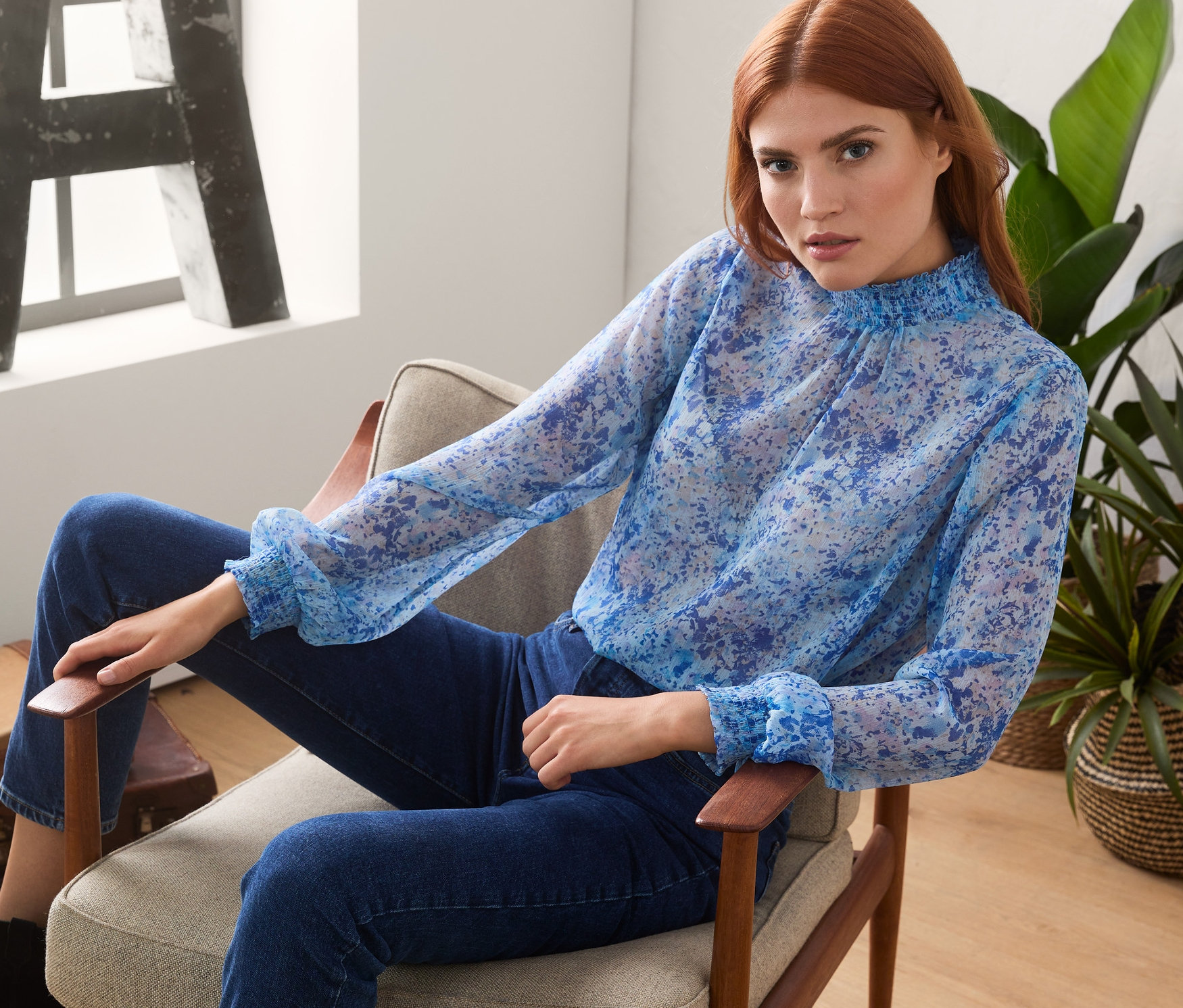 Frau sitzt im Sessel und trägt eine Bluse mit Stehkragen.