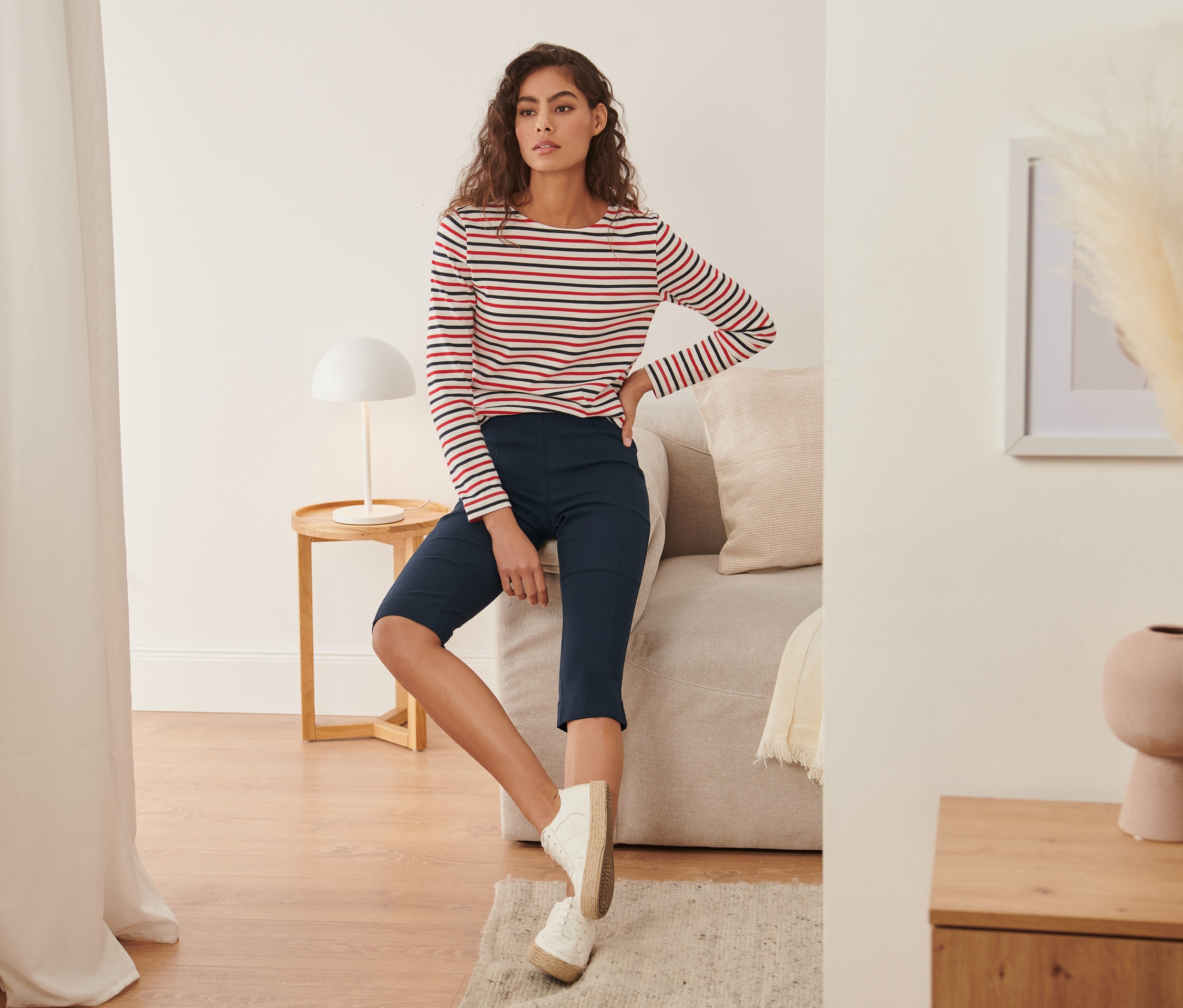 Frau sitzt auf Sofa und trägt eine 3/4-Stretchhose und ein gestreiftes Oberteil.