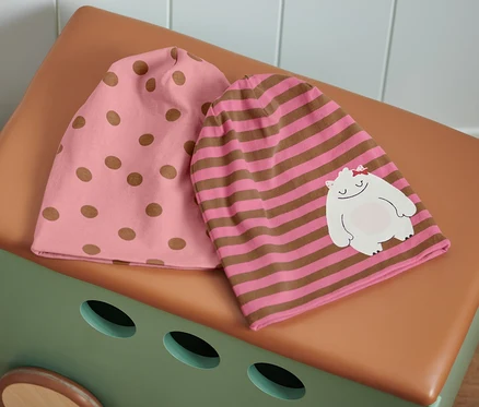 Zwei rosa und braune Mützen liegen auf einer braun-beigen Spielzeugkiste: eine rosa Mütze mit braunen Punkten und eine rosa-braun gestreifte Mütze mit einem Aufnäher eines weißen Monsters.