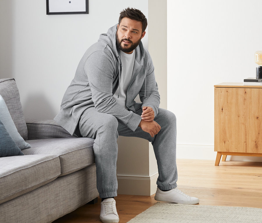 Ein Mann sitzt auf einem Sofa und trägt eine graue Sweatjacke, eine graue Sweathose und ein weißes T-Shirt mit Rundhalsausschnitt. Er trägt weiße Turnschuhe. Im Hintergrund ist eine Kommode zu sehen. Außerdem trägt er 4 Paar Merino-Socken, schwarz, dunkelblau, anthrazit und grau.
