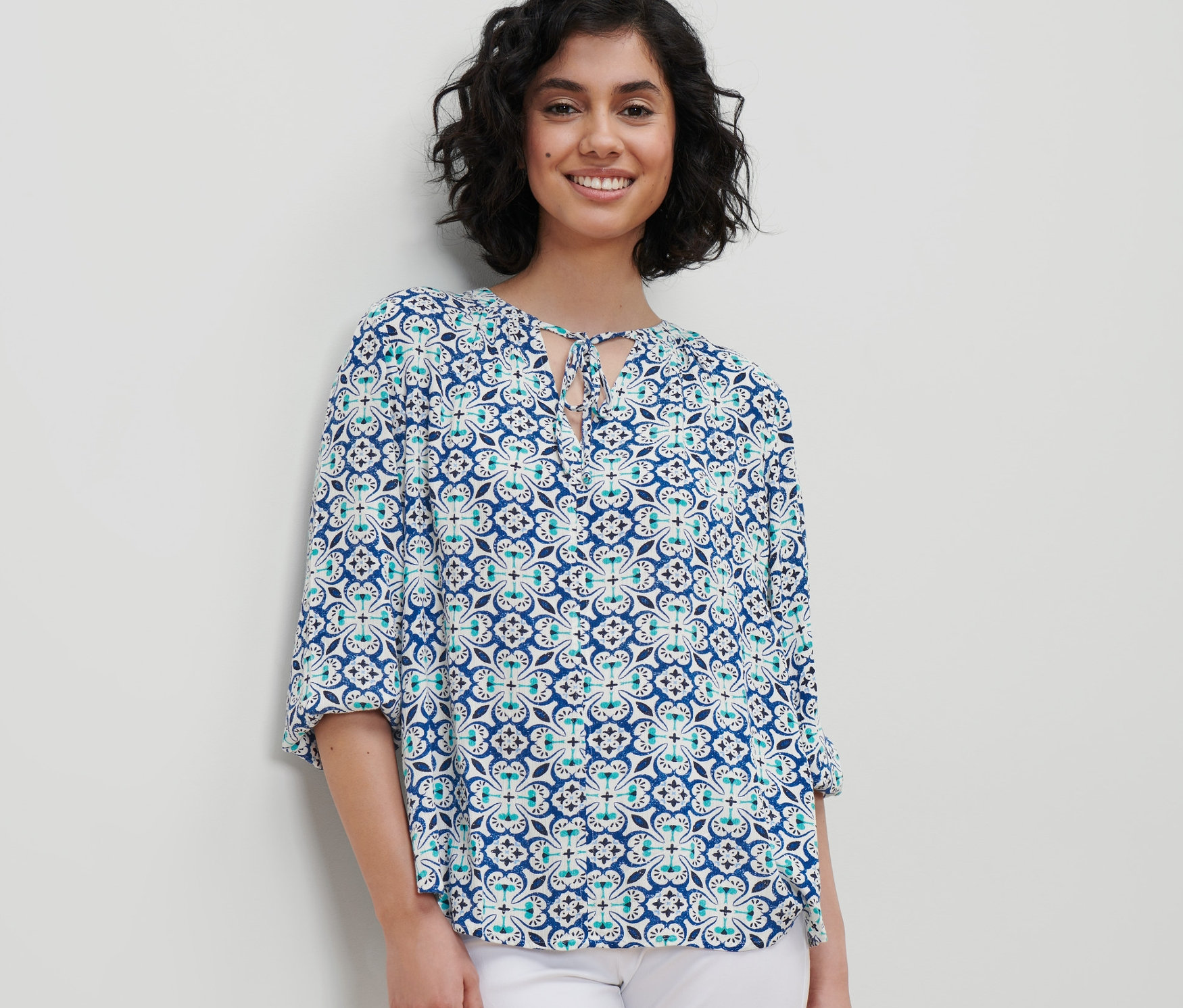 Frau trägt eine Tunika-Bluse mit Alloverprint und weiße Hose.