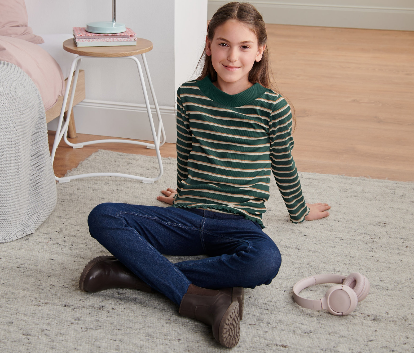Mädchen sitzt auf Teppich und trägt ein geringeltes, grünes Rippshirt, Jeans und Stiefel.