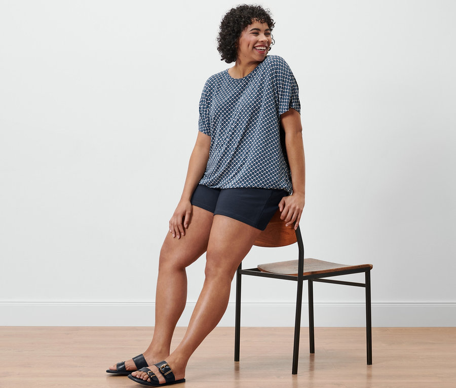 Frau sitzt seitlich auf einem Stuhl, trägt ein bedrucktes Blusenshirt und navy Sweatshorts.