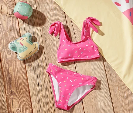 Auf einem Holzboden liegen ein Kinder-Bikini, ein Eiscreme-Strandtuch und Spielzeug.