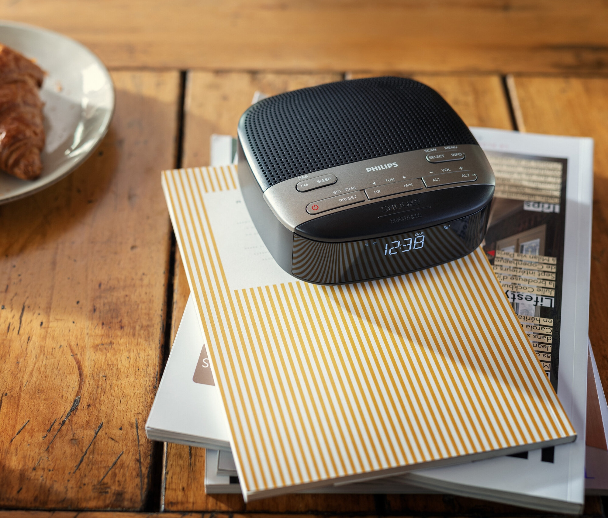 Auf einem Holztisch liegt ein Philips Radiowecker »TAR3505« auf einem Stapel Zeitschriften neben einem Teller mit Croissants und einer Tasse Tee.
