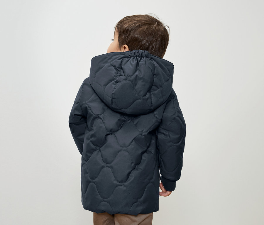 Junge mit dunkelblauer Steppjacke mit Kapuze, der mit dem Rücken zur Kamera steht.
