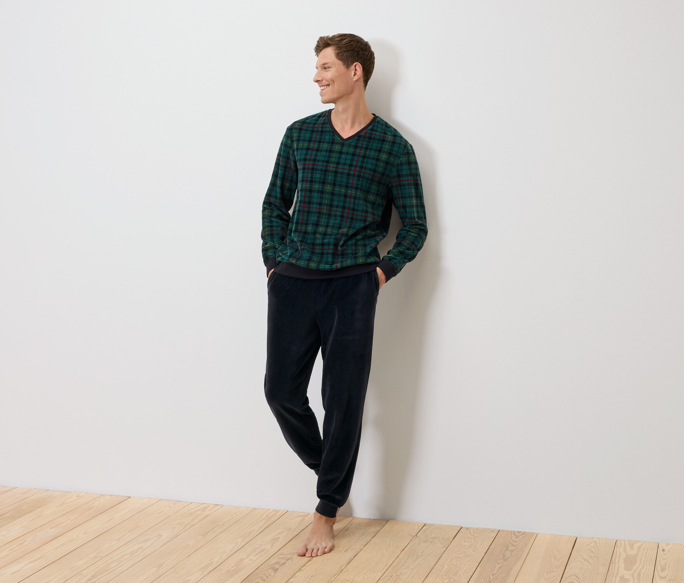 Ein Mann steht an einer Wand, bekleidet mit einem grün karierten Pullover und dunkelblauen Samthosen.