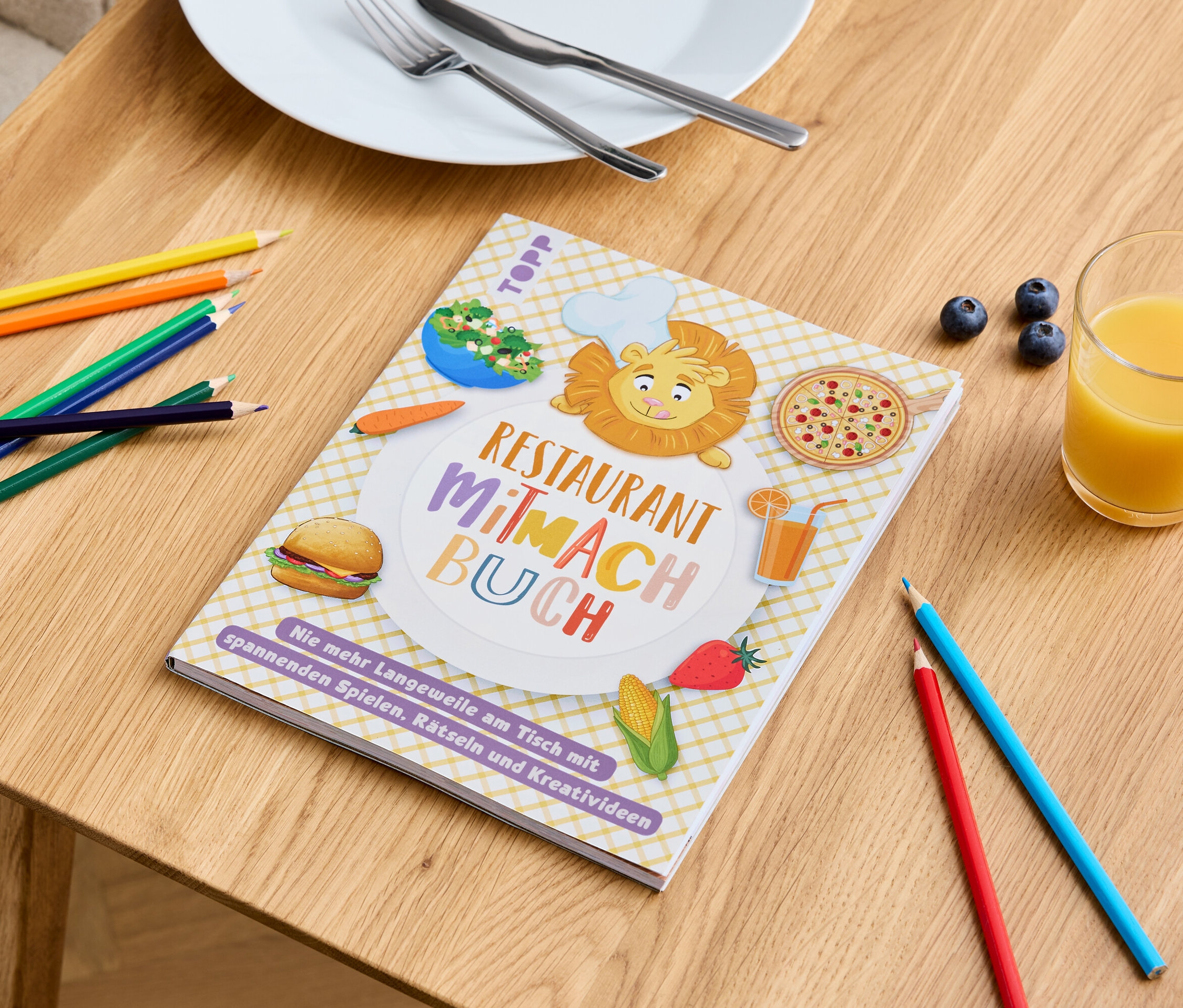 Auf einem Tisch liegen ein "Restaurant Mitmach Buch" mit Essensbildern, Buntstifte, ein Glas Saft und ein Teller mit Besteck.