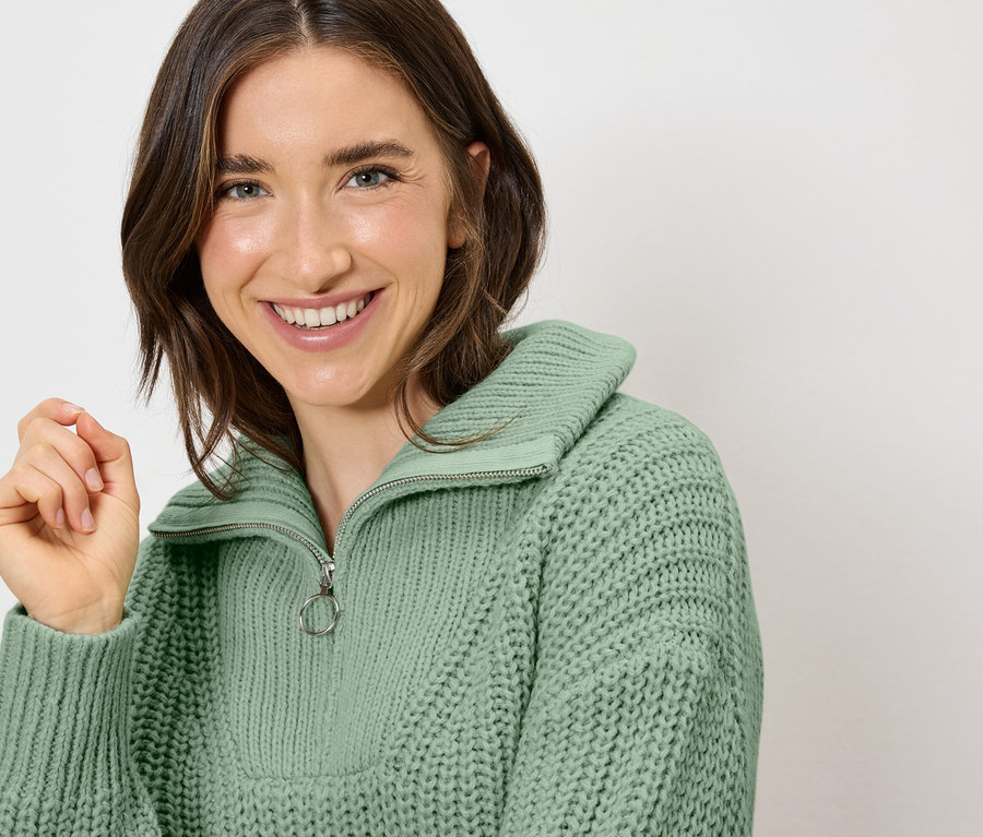 Eine lächelnde Frau trägt einen hellgrünen Strickpullover mit Reißverschluss.