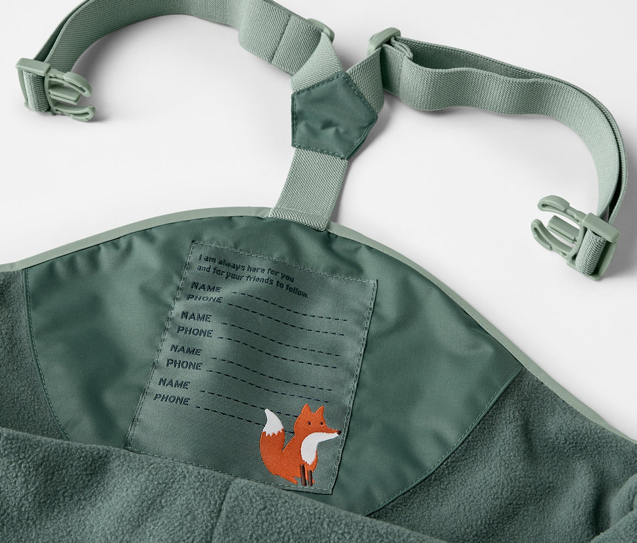 Detail einer grünen Kinder-Thermo-Regenhose mit Hosenträgern und Namensschild mit Fuchsmotiv.