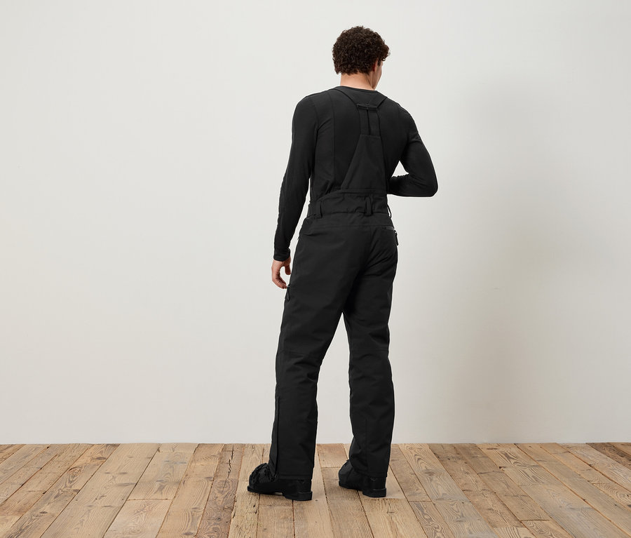 Ein Mann mit dem Rücken zur Kamera trägt eine schwarze Latzhose und ein schwarzes Langarmshirt.