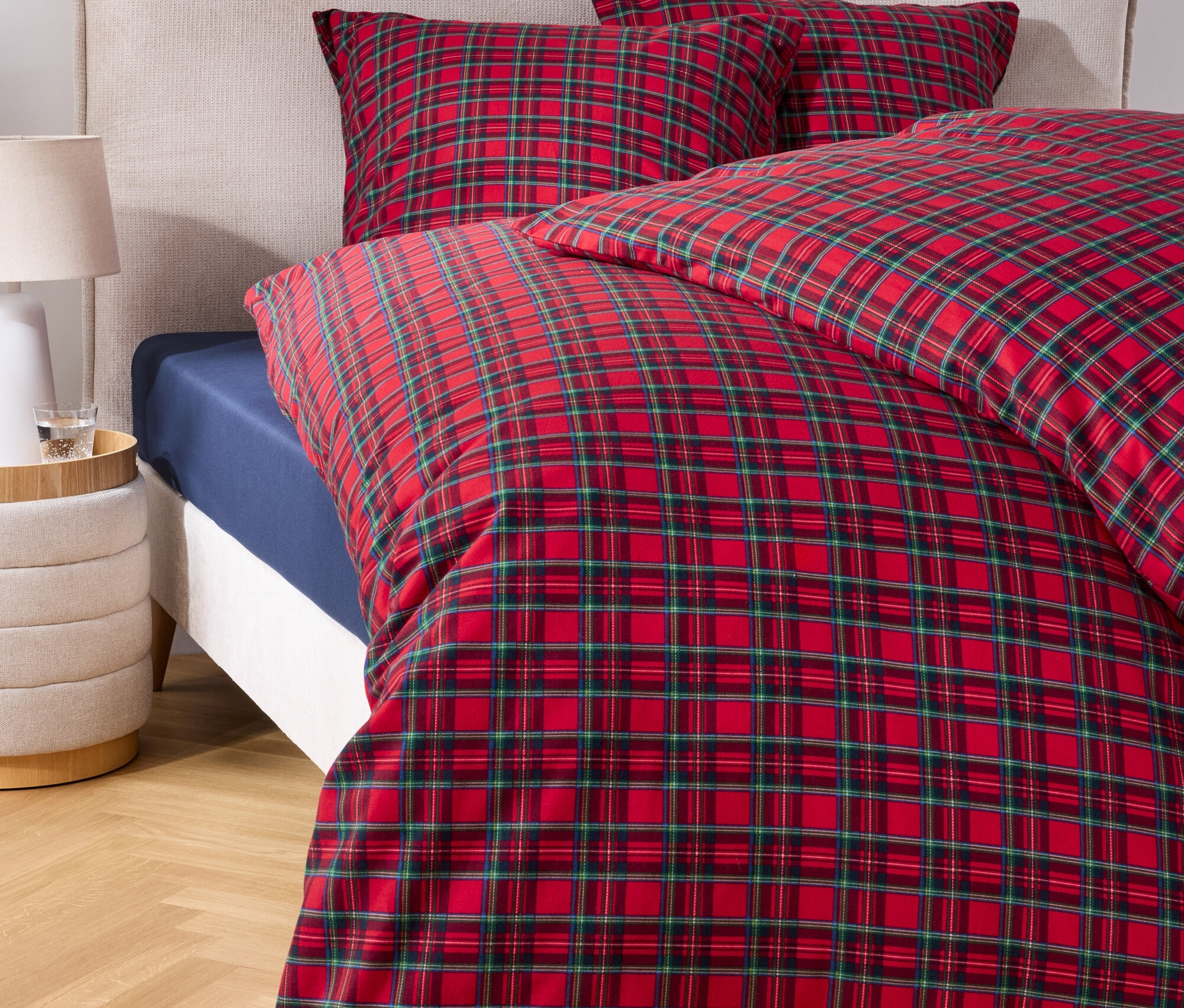 Rot-grün karierte Bettdecke auf einem Bett mit zwei Kissen und einem Nachttisch.