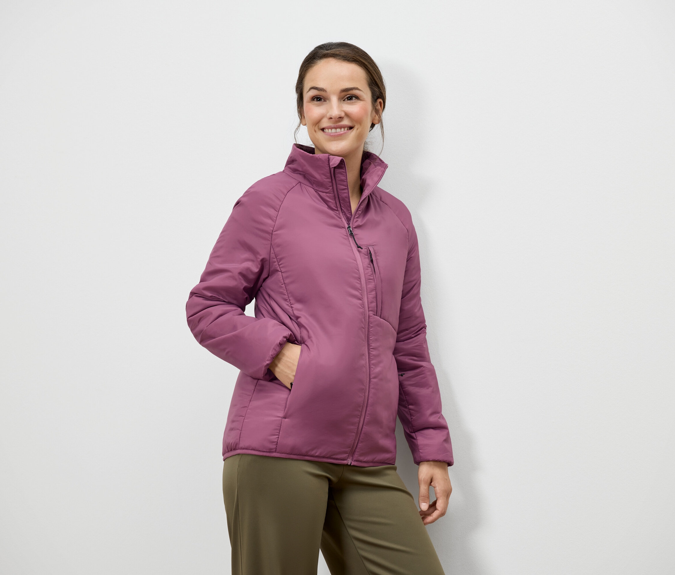 Frau mit braunen Haaren und einem Lächeln posiert in einer pinkfarbenen Jacke und grünen Hosen vor einer weißen Wand.