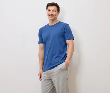 Lächelnder Mann in blauem T-Shirt und grauer Jogginghose posiert vor weißem Hintergrund.