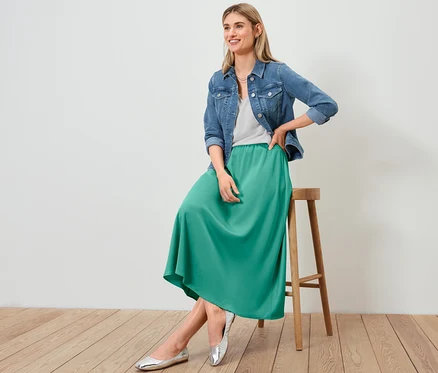 Frau sitzt auf einem Hocker und trägt eine Jeansjacke, einen Satinrock und ein strukturiertes Shirt in Weiß.
