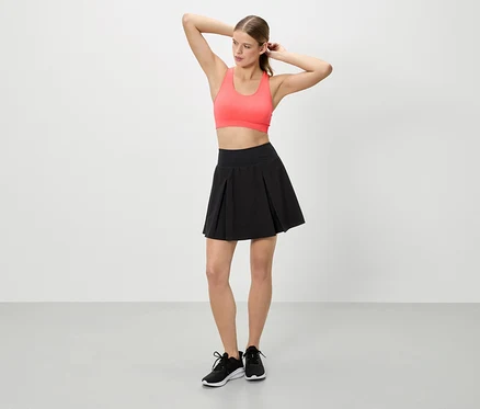 Frau mit erhobenen Händen trägt ein korallfarbenes Sportoberteil, einen schwarzen Rock und schwarze Turnschuhe.