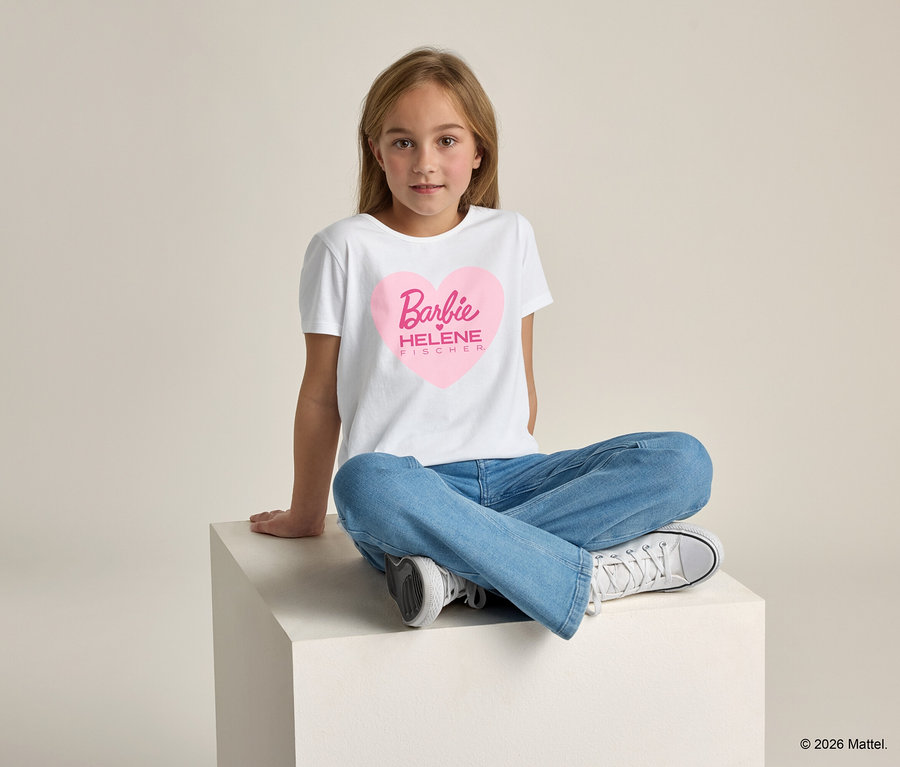Mädchen sitzt mit gekreuzten Beinen auf einem weißen Würfel und trägt ein weißes T-Shirt mit einem pinkfarbenen Herz, in dem „Barbie HELENE FISCHER“ steht.