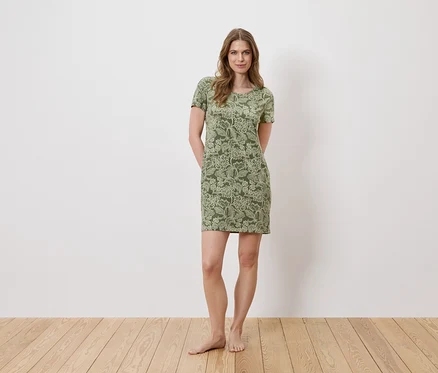 Eine Frau steht barfuß vor einer weißen Wand und trägt ein grünes, kurzärmeliges Kleid mit Blumenmuster.