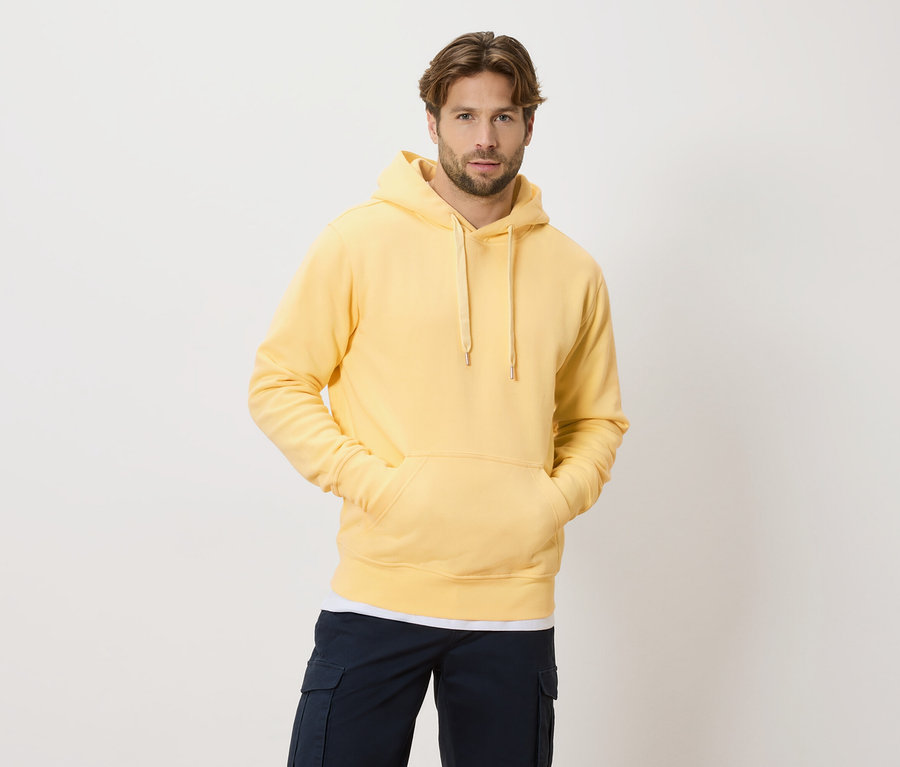 Mann mit hellbraunen Haaren und Bart trägt einen gelben Hoodie und marineblaue Cargohosen vor einem weißen Hintergrund.