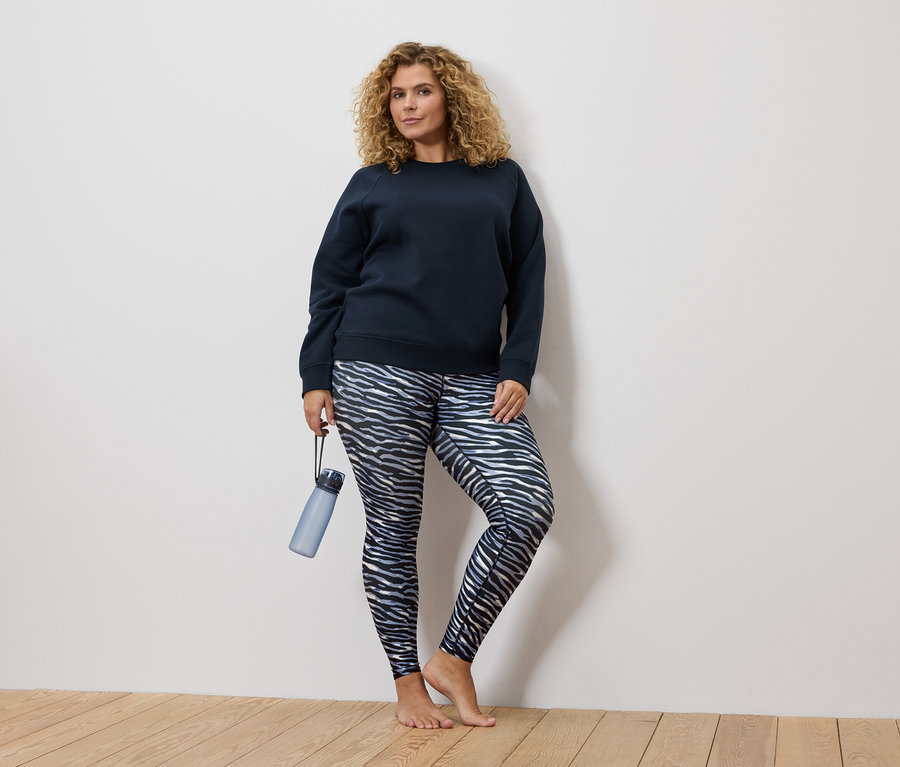 Frau mit blonden Locken trägt einen marineblauen Pullover und Leggings mit Zebraprint. Sie hält eine blaue Wasserflasche.