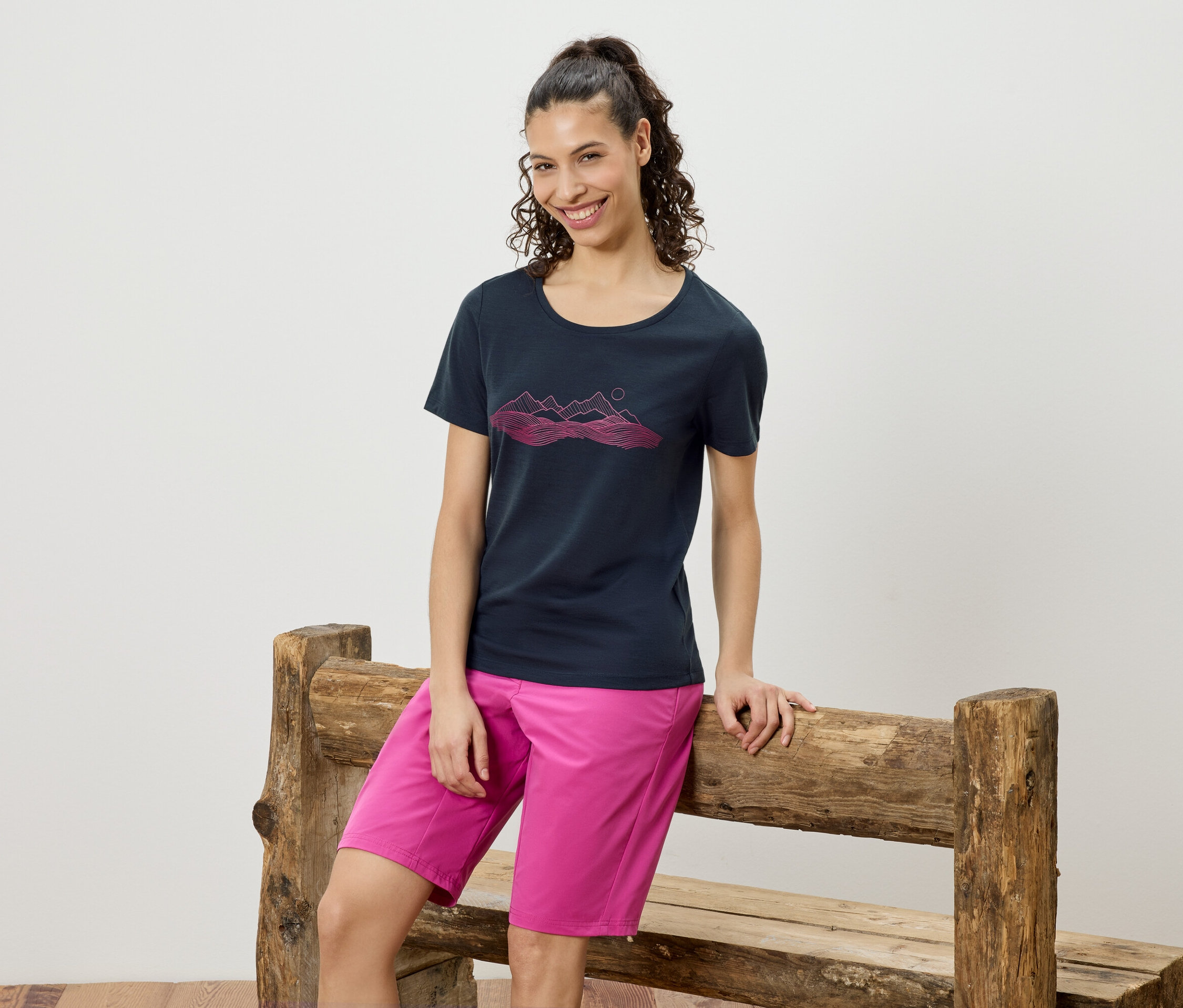 Frau lehnt an einer Holzbank, trägt ein marineblaues T-Shirt mit einem pinkfarbenen Bergmotiv und pinkfarbene Shorts.