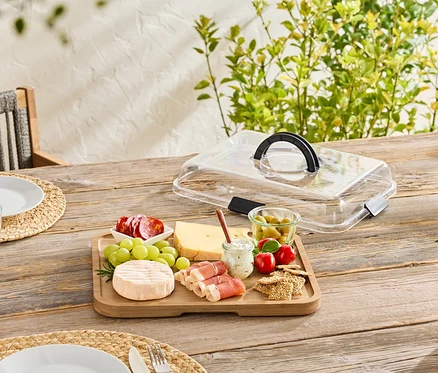 Auf einem Holztisch steht ein Holzbrett mit Käse, Wurst, Oliven und Cracker, teilweise mit einer transparenten Abdeckung bedeckt.