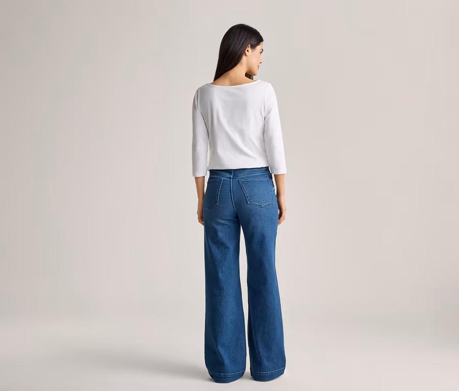 Rückansicht einer Frau mit dunklen Haaren, die ein weißes, langärmeliges Oberteil und eine weite, blaue Jeans trägt.