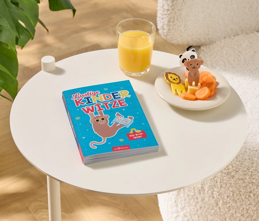 Auf einem Tisch liegen ein Buch mit dem Titel "Lustige Kinderwitze", ein Glas Orangensaft und ein Teller mit Karotten und Tierfiguren.