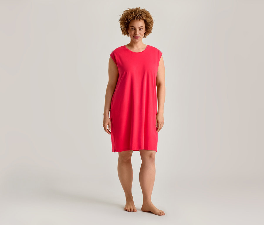 Eine Frau mit lockigem Haar steht barfuß und trägt ein kurzes, locker sitzendes, leuchtend pinkes Kleid.