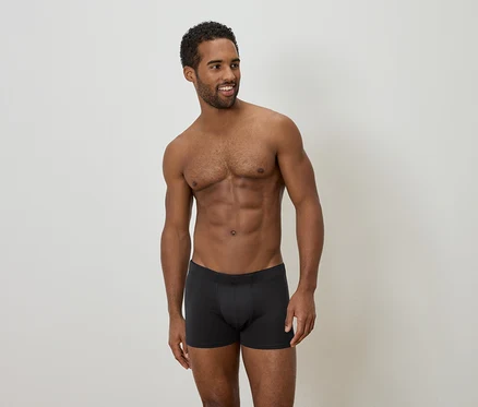Ein Mann mit dunkler Hautfarbe und dunklen, lockigen Haaren steht vor einem neutralen Hintergrund und trägt schwarze Boxershorts.