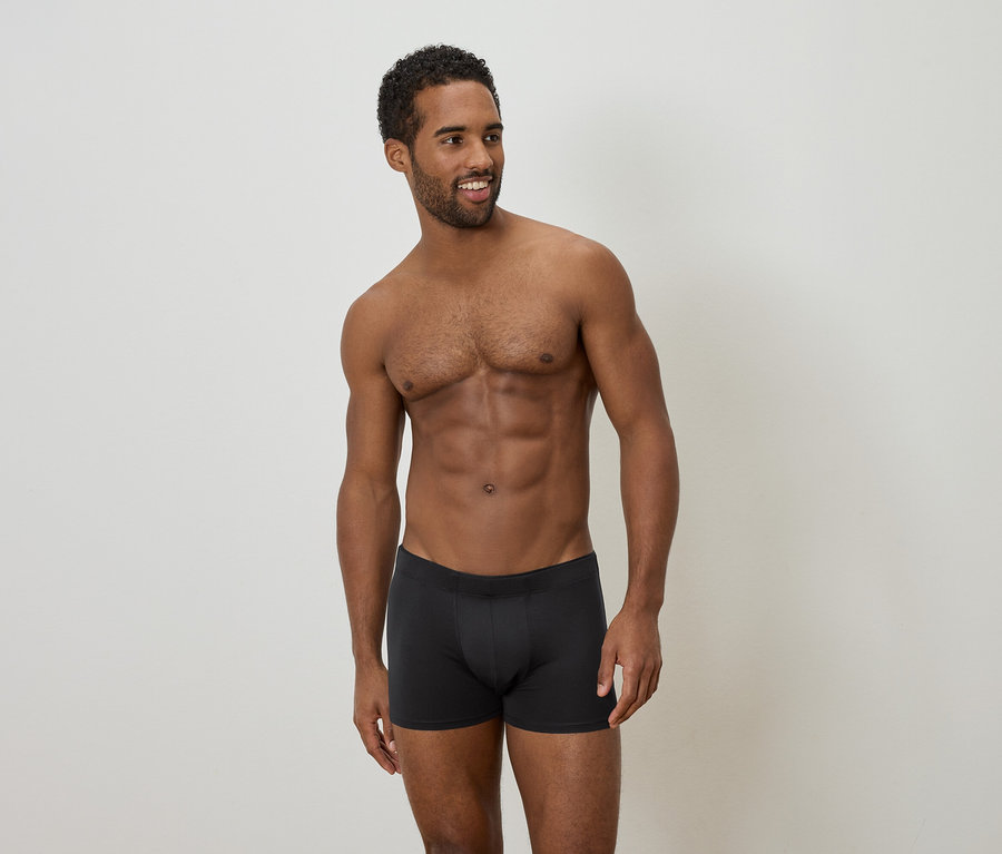 Ein Mann mit dunkler Hautfarbe und dunklen, lockigen Haaren steht vor einem neutralen Hintergrund und trägt schwarze Boxershorts.