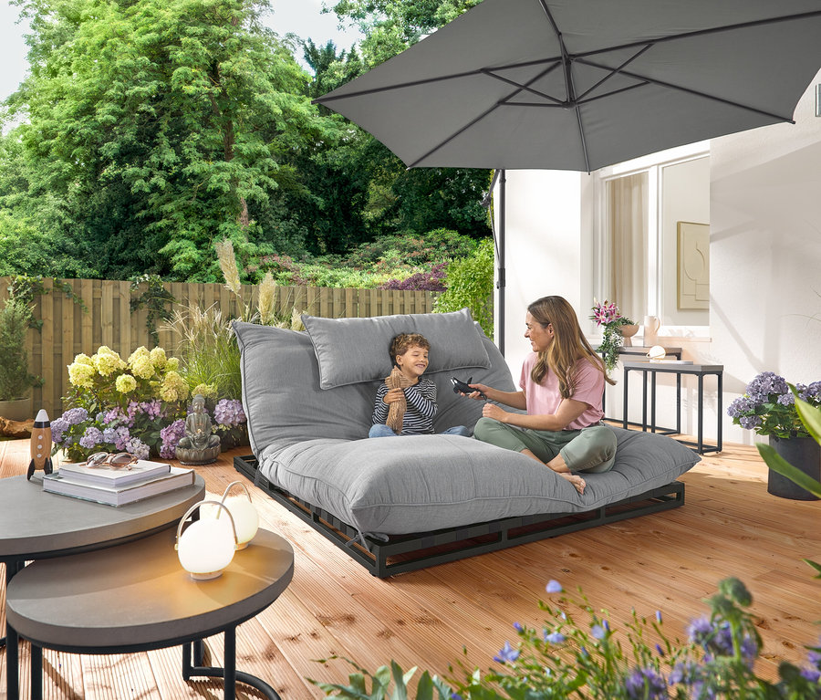 Eine Frau und ein Kind sitzen auf einer Premium-Doppel-Gartenliege mit Creacryl®-Allwetterstoff. Im Garten stehen 2 Garten-Couchtische und 2 Garten-Konsolentische mit Polyzement-Tischplatten und 3 Akku-Tischleuchten.