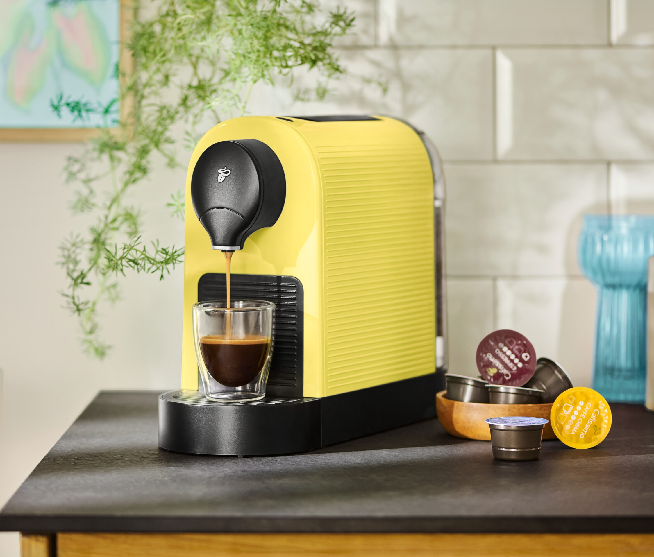 Eine gelbe Kaffeemaschine füllt eine Tasse mit Kaffee, Kapseln liegen daneben.