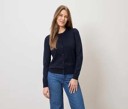Frau mit langen braunen Haaren, trägt eine marineblaue Strickjacke mit Knöpfen und blaue Jeans vor einem hellen Hintergrund.