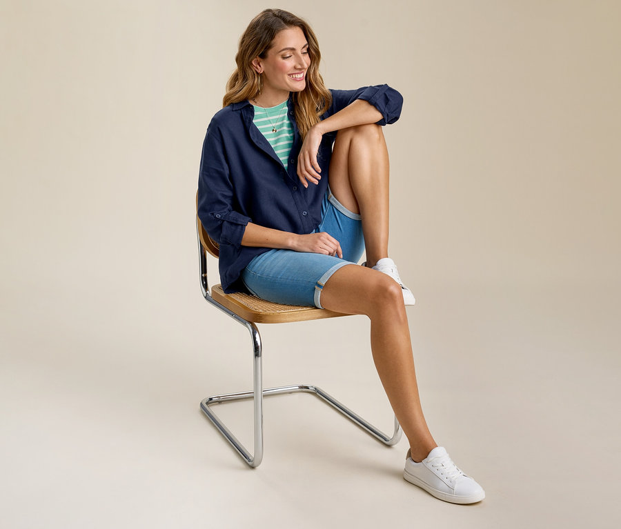 Frau sitzt auf Stuhl und trägt ein mint-weiß gestreiftes Basic-T-Shirt unter einem marineblauen Hemd, Jeansshorts und weiße Turnschuhe.