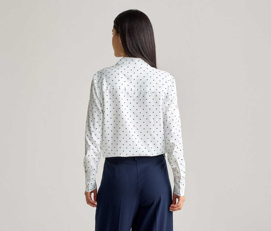 Rückansicht einer Frau mit langen, dunklen Haaren, die eine weiße, langärmlige Bluse mit schwarzen Punkten und eine weite, marineblaue Hose trägt.