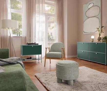 Wohnzimmer mit grünem Sofa, einer Eglo Stehleuchte »Fondachelli«, einem Sideboard Metall »CN3« mit 1 Klappenfach und Plattenspieler. Ein Sessel mit Holzgestell und ein Sitzpouf stehen auf einem Teppich. Rechts ein Sideboard Metall »CN3« mit 4 Klappenfächern und einer Trio Keramik-Tischleuchte.