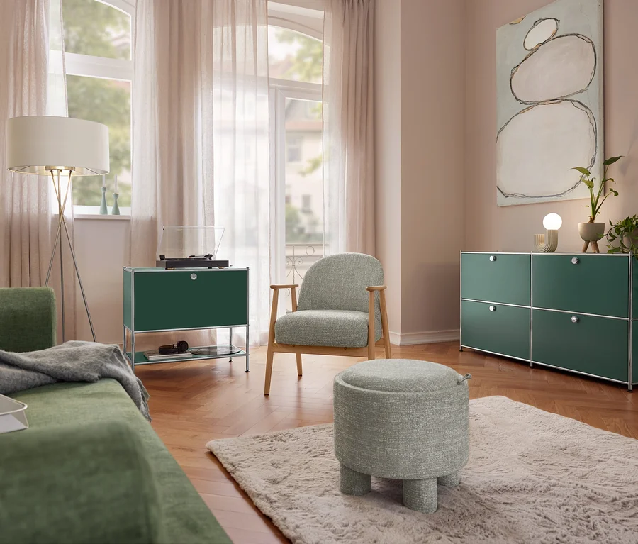 Wohnzimmer mit grünem Sofa, einer Eglo Stehleuchte »Fondachelli«, einem Sideboard Metall »CN3« mit 1 Klappenfach und Plattenspieler. Ein Sessel mit Holzgestell und ein Sitzpouf stehen auf einem Teppich. Rechts ein Sideboard Metall »CN3« mit 4 Klappenfächern und einer Trio Keramik-Tischleuchte.