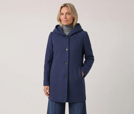 Frau in einem blauen Kapuzenmantel über einem grauen Rollkragenpullover, mit der linken Hand in der Manteltasche, blickt nach vorne.