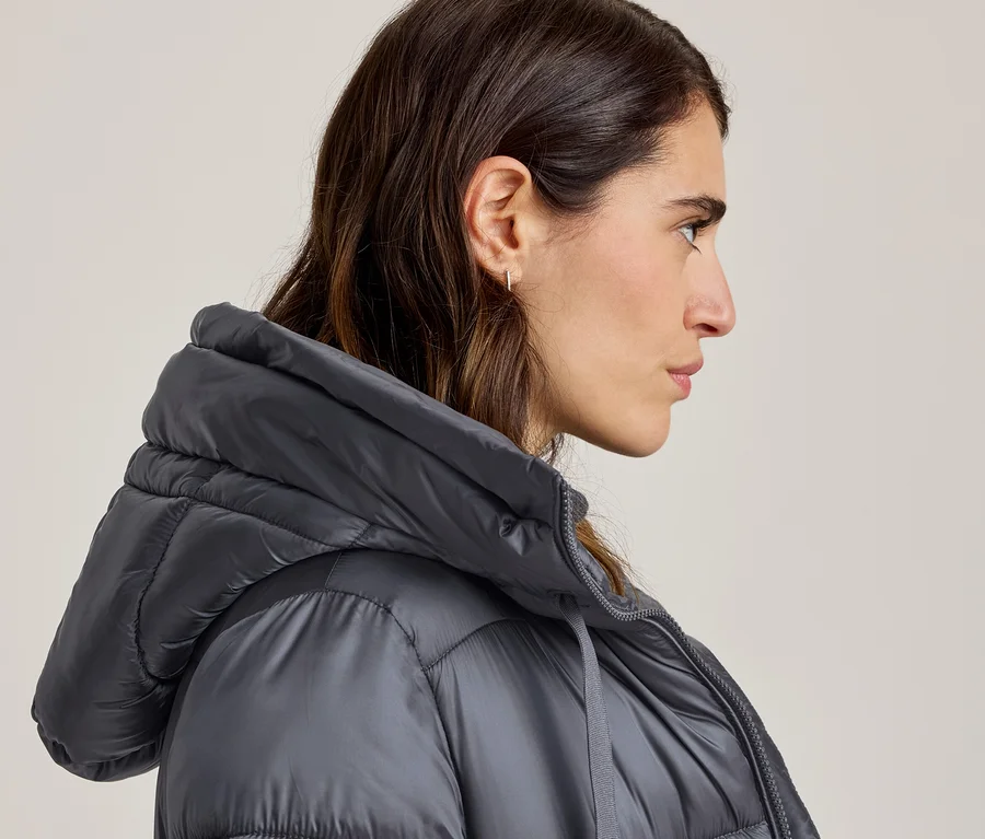 Frau im Profil trägt eine graue Steppjacke mit Kapuze, ihr dunkles Haar und ein Ohrring sind sichtbar.
