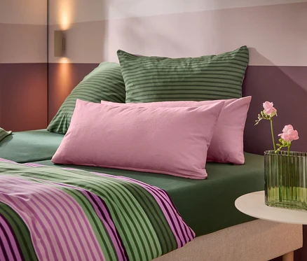 Ein Bett mit grünem Spannbetttuch, grün gestreiften Kissen, rosa Kissen und einer gestreiften Bettdecke. Ein weißer Beistelltisch trägt eine grüne gerippte Vase mit rosa Blumen. Die Wand dahinter hat horizontale rosa und lila Streifen mit zwei Wandleuchten.